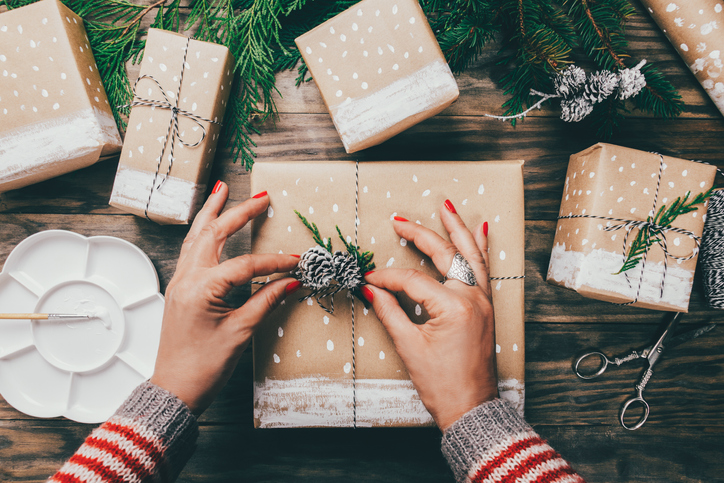Woman´s hands wrapping Christmas presents on brown paper decorated with painted snow, fir branches and pinecones on a rustic wooden board.