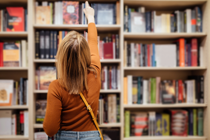 Pretty young girl with loose long blondie hair standing and holding an open book between book shelves in the library, reading and looking into the book